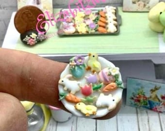 May include: A white plate with a variety of miniature Easter-themed cookies, including bunnies, chicks, carrots, and eggs. The cookies are decorated with pastel colors and icing.