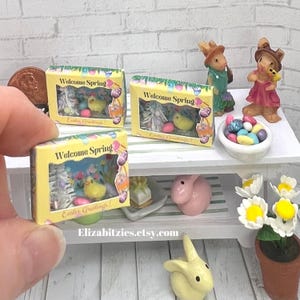 May include: Three miniature boxes with a yellow background and a clear window showing a miniature Easter scene. The boxes say "Welcome Spring" and "Easter Greetings!"  The boxes are on a white shelf with miniature Easter decorations.