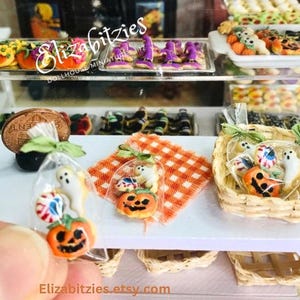 May include: A miniature Halloween bakery display with a variety of cookies in bags and baskets. The cookies are decorated with ghosts, pumpkins, and candy corn. The display is set up on a shelf with a checkered tablecloth.