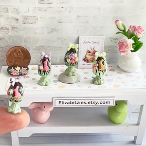 May include: A miniature white table with a green and pink Easter egg decoration on top. The table has a sign that reads "Elizabitzzies.etsy.com". There are four other Easter egg decorations on the table, each with a different design.