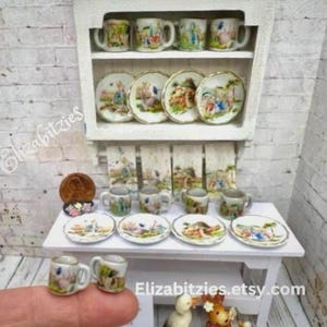 May include: A miniature dollhouse kitchen set with a white cabinet, a white table, and a set of miniature teacups and saucers with a Peter Rabbit theme. The teacups and saucers are decorated with images of Peter Rabbit and other characters from the book.