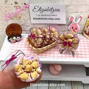 May include: A miniature display of bunny-shaped cookies in clear plastic containers with pink bows. The cookies are arranged in a wicker basket and on a white table with a pink and white checkered tablecloth. The text "Elizabitzzies 1:12 Miniatures Elizabitzzies.etsy.com" is visible on a sign.