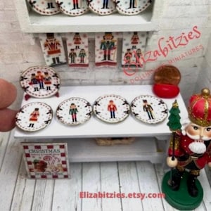 May include: A set of six miniature porcelain plates featuring a nutcracker design. The plates are white with red and gold accents. The plates are arranged on a white shelf with a miniature nutcracker figurine and a Christmas-themed book.