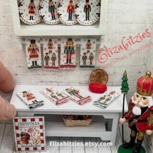 May include: Miniature Christmas-themed dollhouse decor featuring a white table with nutcracker-themed towels and plates. A miniature nutcracker soldier stands nearby. A sign reads "Christmas" with candy cane and gingerbread designs. The Etsy shop name is visible.