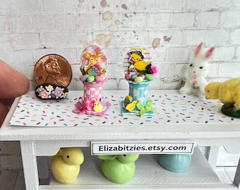May include: A white table with a pink and blue confetti patterned surface. Two miniature Easter baskets with pastel colored chicks and bunnies are on the table.  Three pastel colored bunny figurines are on the bottom shelf. The text "Elizabitzzies.etsy.com" is on the table.