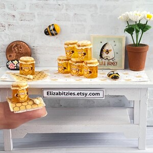 May include: A miniature white table with a sign that reads "Elizabitzzies.etsy.com" and a stack of miniature honey jars with a bee design. The table is decorated with a white tablecloth and a small white flower arrangement in a terracotta pot.