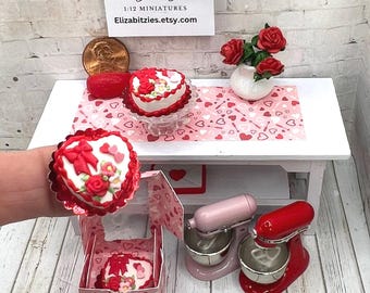May include: A miniature kitchen set with a pink and red stand mixer, a heart-shaped cake with red and white frosting, and a box of heart-shaped cookies. The set is decorated with a red and white heart-shaped tablecloth and a vase of red roses.