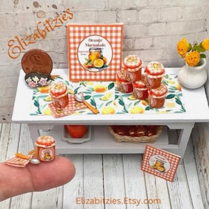 May include: A miniature dollhouse kitchen scene with a white table, orange marmalade jars, a spoon, and a small basket of oranges. The table has a checkered orange and white tablecloth with a lemon and orange design. The jars are labeled "Orange Marmalade".