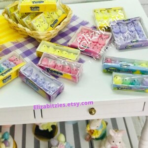 May include: A collection of miniature boxes of Peeps marshmallow candies in various colors, including yellow, pink, purple, and green. The boxes are arranged on a white surface with a purple and white checkered tablecloth in the background.
