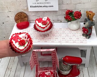 May include: A miniature dollhouse scene with a red and white heart-shaped cake on a glass pedestal, a red and white heart-shaped cake in a pink and white box, a red stand mixer, and a white vase with red roses.