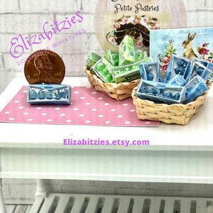 May include: A miniature dollhouse scene with two wicker baskets filled with miniature boxes of pastel green and blue eggs. The baskets are on a white table with a pink polka dot tablecloth. The text "Petite Pastries" is visible on a sign behind the baskets.