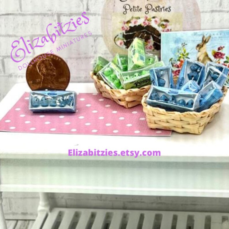 May include: A miniature dollhouse scene with two wicker baskets filled with miniature boxes of pastel green and blue eggs. The baskets are on a white table with a pink polka dot tablecloth. The text "Petite Pastries" is visible on a sign behind the baskets.