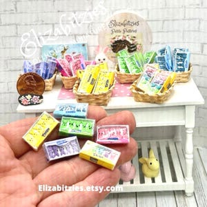 May include: A miniature dollhouse display of Peeps marshmallow candies in various colors, including pink, yellow, blue, and green. The candies are arranged in small wicker baskets on a white table with a pink and white checkered tablecloth. The candies are packaged in boxes with the word "Peeps" printed on them. The display is set up for a dollhouse or miniature scene.