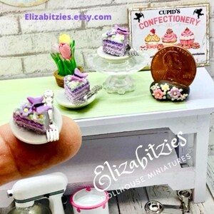May include: Three miniature purple cakes with butterfly decorations on white plates. The cakes are on a white table with a green tablecloth. A sign that says "Cupid's Confectionery" is in the background.