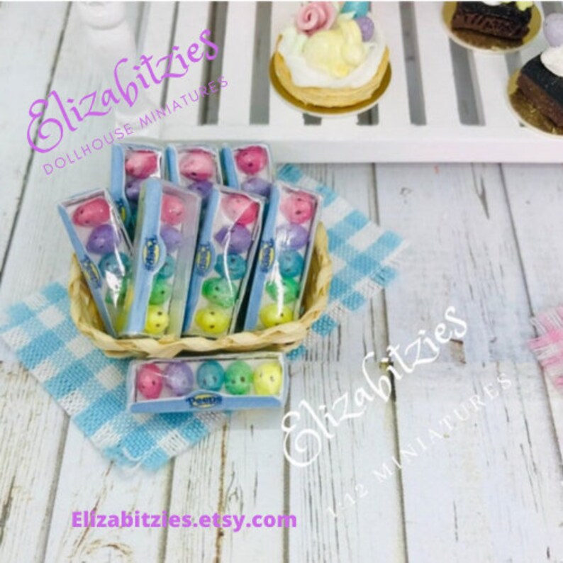 May include: A basket filled with miniature boxes of pastel-colored candy eggs. The boxes are labeled "Peeps" and are arranged in a decorative display. The basket is on a blue and white checkered surface. The image includes the text "Elizabitzzies Dollhouse Miniatures" and "Elizabitzzies 1:12 Miniatures".