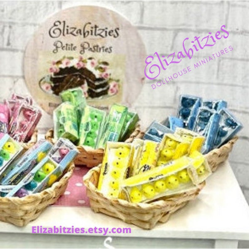 May include: A collection of miniature candy boxes in wicker baskets. The boxes are filled with colorful candies, including yellow, green, and blue. The boxes are arranged on a white surface with a sign that reads "Elizabitties Petite Pastries".