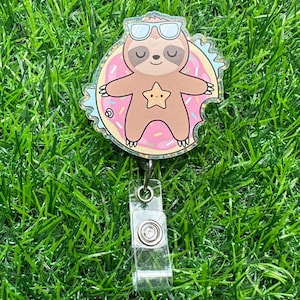 May include: A pink and blue glitter badge reel with a cartoon sloth wearing sunglasses and lying on a donut.