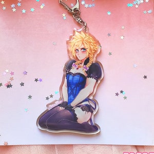 May include: A clear acrylic keychain featuring an anime-style character with blonde hair, wearing a blue and black corset and stockings. The character is sitting with their legs crossed and has a serious expression. The keychain is on a pink background with silver glitter stars.