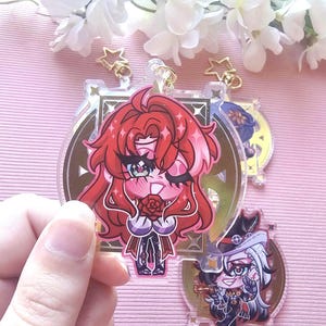 May include: Clear acrylic charms with anime-style characters in bright colours. The charms have gold-coloured hardware and detailed illustrations. The text "EDENFIRE" is visible on one of the charms.