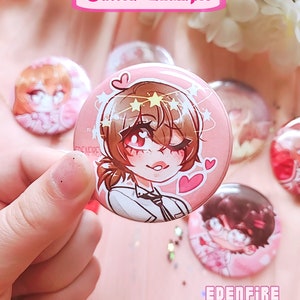 Op de afbeelding: Een roze knoop met een cartoonillustratie van een persoon met bruin haar, die een wit shirt en een zwarte stropdas draagt. De persoon heeft sterren en harten rond zijn hoofd. De knoop heeft de tekst "EDENFIRE" erop.