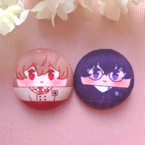 May include: Two circular plush badges featuring anime-style character faces. The left badge shows a character with light brown hair and a white uniform. The right badge shows a character with dark hair, glasses, and a dark uniform. Text reads EDENFIRE.