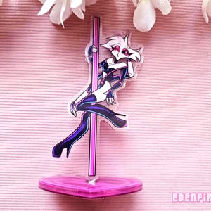 Könnte beinhalten: Ein transparenter Acryl-Ständer mit einer rosa Basis. Der Ständer zeigt eine Cartoon-Illustration einer Figur mit weißen Haaren und schwarzer Kleidung, die eine rosa Stange hält.