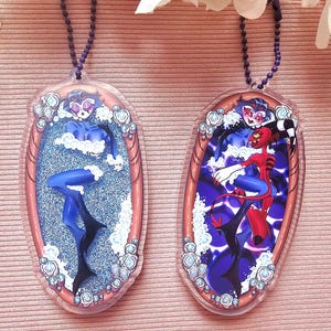 May include: Two oval-shaped acrylic keychains with cartoon character designs. One features a blue figure in a blue glitter background, the other a red and blue character. Both have a purple chain and the text "EDENFIRE".