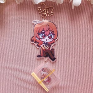 Peut inclure: Un porte-clés en acrylique transparent représentant un personnage de dessin animé aux cheveux et aux yeux rouges, vêtu d'un costume marron. Un petit récipient de nourriture avec des baguettes est attaché en dessous. Le porte-clés est suspendu à un fermoir doré en forme de nœud. Le texte "EDENFIRE" est visible.