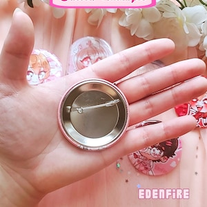 Op de afbeelding: Een close-up van een roze knoop met een metalen achterkant. De knoop heeft een cartoonillustratie van een personage met zwart haar en een bril. De tekst "Edenfire" is zichtbaar op de knoop.