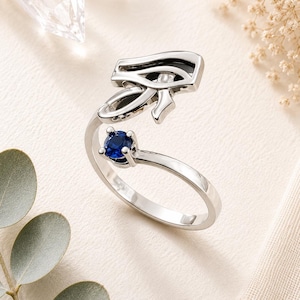 May include: A silver-toned open ring featuring a polished Eye of Horus symbol on one end and a single, round, deep blue gemstone set in a four-prong mount on the other end. The band is smooth and metallic, designed with an adjustable open gap.