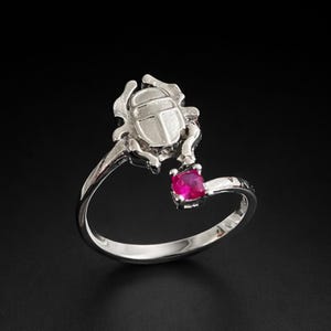 May include: A silver ring featuring a scarab beetle design with a ruby-colored gemstone. The ring has a polished finish and a delicate band. The scarab beetle is a prominent detail, adding a unique touch to the jewelry.