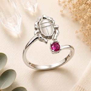 Pode incluir: Um anel aberto de tom prateado com um motivo de escaravelho esculpido numa extremidade e uma única pedra preciosa redonda de cor rosa fúcsia vibrante, fixada com quatro garras na outra extremidade, com um acabamento em metal polido.