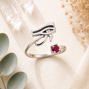 May include: A silver-toned open ring featuring the Eye of Horus symbol on one end and a small, round, vibrant red gemstone set in a four-prong mount on the other end. The band is smooth and polished, creating a bypass style design.