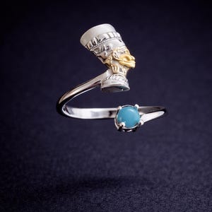 Puede incluir: Anillo de plata con un busto de Nefertiti con detalles dorados y una piedra turquesa. El diseño del anillo está inspirado en el arte egipcio antiguo, con una representación detallada del tocado y el perfil de la reina. La piedra es redonda y de color azul claro.