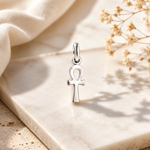 May include: A polished silver ankh pendant featuring a classic looped top and a vertical stem, suspended from a small circular jump ring. The metallic surface reflects light, highlighting the smooth, clean lines of this ancient Egyptian symbol.