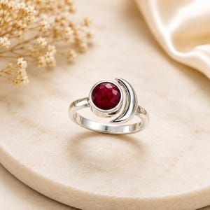 May include: A silver ring featuring a round, faceted deep red gemstone set in a bezel, accompanied by a polished silver crescent moon shape that curves alongside the stone setting, creating a celestial-inspired design on a smooth metal band.