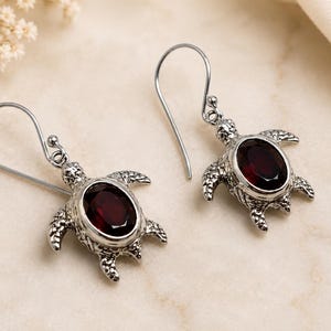 May include: A pair of silver-toned drop earrings featuring a turtle-shaped design. Each turtle has a textured metal shell and a central oval-shaped deep red gemstone. The earrings are attached to simple silver-toned ear wires for hanging.