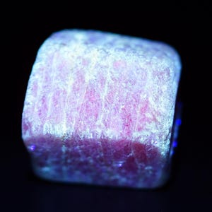 Może przedstawiać: Zbliżenie różowo-białego okazu mineralnego na czarnym tle. Minerał ma szorstką, teksturowaną powierzchnię z widocznymi strukturami krystalicznymi. Krawędzie minerału świecą niebieskim odcieniem.
