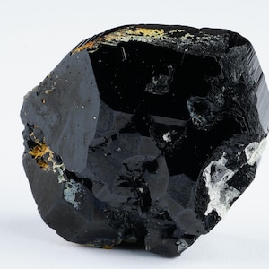 May include: A large, faceted black crystal with a glossy surface. The crystal has a complex, irregular shape with areas of gold and white mineral deposits. The background is plain white, highlighting the dark, reflective qualities of the stone.