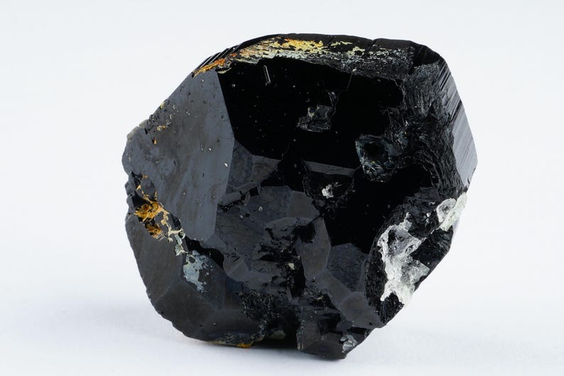 May include: A large, faceted black crystal with a glossy surface. The crystal has a complex, irregular shape with areas of gold and white mineral deposits. The background is plain white, highlighting the dark, reflective qualities of the stone.