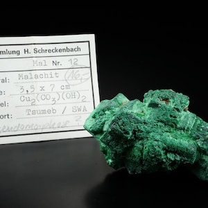 Op de afbeelding: Een groen malachiet mineraal specimen met een ruw, getextureerd oppervlak. Het specimen is gelabeld met een witte kaart waarop "Sammlung H. Schreckenbach" staat en details over het mineraal bevat, waaronder de grootte, chemische formule en herkomst. Het label geeft aan dat het mineraal 3,5 x 7 cm groot is en afkomstig is uit Tsumeb, SWA.