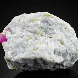 Könnte beinhalten: Nahaufnahme eines rauen, weißen Gesteins mit einem eingebetteten, leuchtend pinkfarbenen Rubinkristall. Das Gestein ist mit kleinen, goldfarbenen Kristallen übersät.
