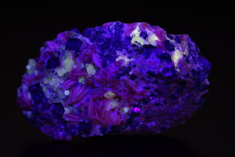 K&ouml;nnte beinhalten: Nahaufnahme eines Mineralproben mit einem Farbschema in Lila, Rosa und Wei&szlig;. Die Probe wird mit UV-Licht beleuchtet, wodurch sie fluoresziert.