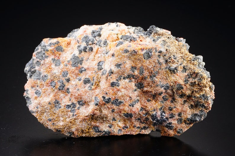 K&ouml;nnte beinhalten: Ein rauer, unregelm&auml;&szlig;ig geformter Stein mit einer rosa-braunen Basis. Der Stein ist mit kleinen, schwarzen, metallisch aussehenden Kristallen bedeckt.