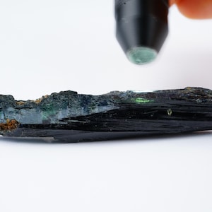May include: A dark green and black crystal with a striated surface. The crystal is about 5 inches long and is resting on a white surface.