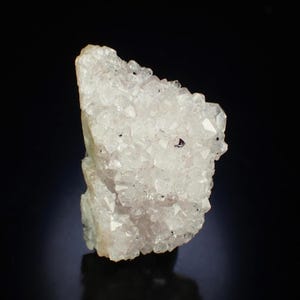 May include: A cluster of clear quartz crystals with a slightly milky appearance. The crystals have sharp, defined points and a rough, textured surface. Small, dark inclusions are scattered throughout the quartz. The background is black.