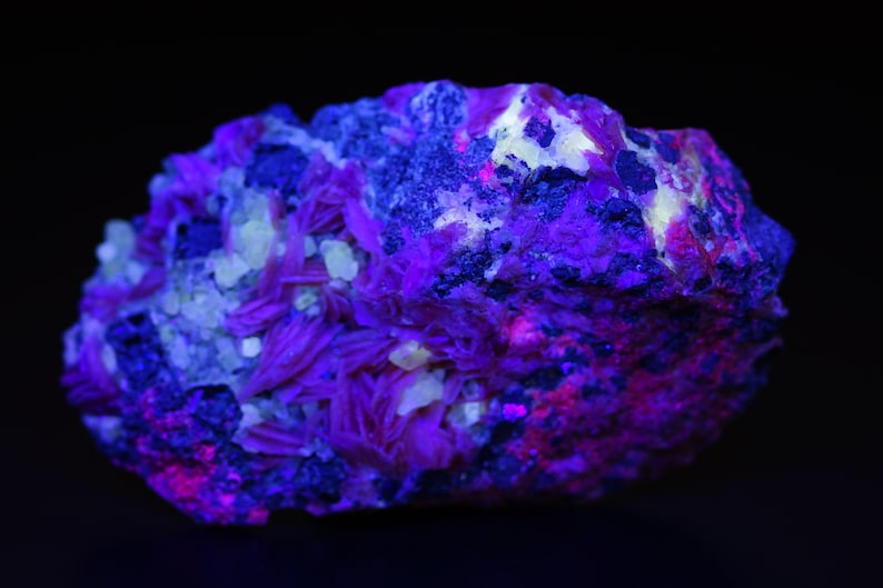 K&ouml;nnte beinhalten: Nahaufnahme eines violetten und rosafarbenen Mineralproben unter UV-Licht. Das Mineral hat eine raue, strukturierte Oberfl&auml;che und ist mit kleinen Kristallen bedeckt.