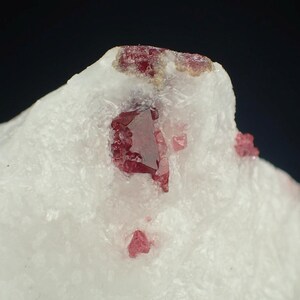 Puede incluir: Primer plano de un espécimen mineral con una matriz blanca con cristales rojos brillantes incrustados. Los cristales son angulares y translúcidos, contrastando con el fondo blanco opaco. La composición resalta la belleza natural de la formación mineral.