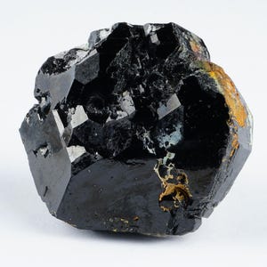 May include: A raw, black tourmaline crystal with a glossy, faceted surface. The gemstone has a complex, irregular shape with areas of crystalline structure and some patches of brown and gold coloration. The background is plain white.