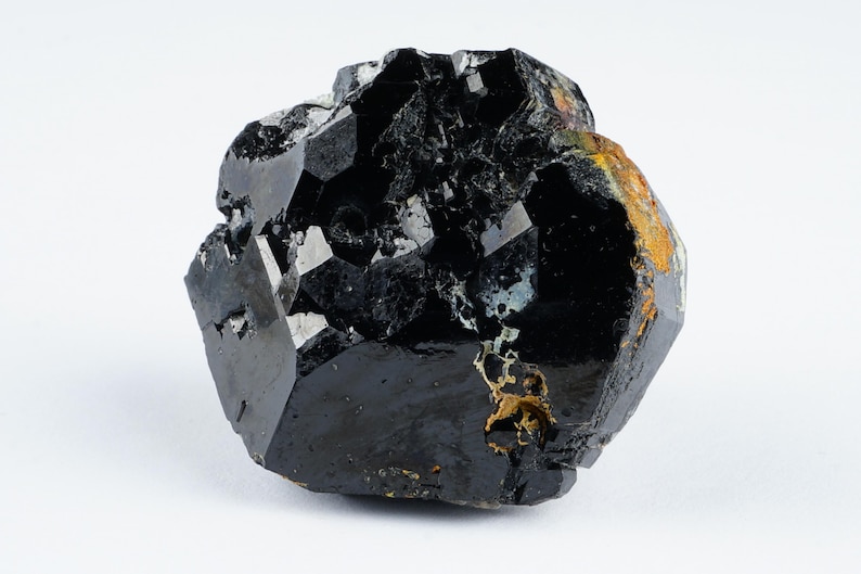 May include: A raw, black tourmaline crystal with a glossy, faceted surface. The gemstone has a complex, irregular shape with areas of crystalline structure and some patches of brown and gold coloration. The background is plain white.